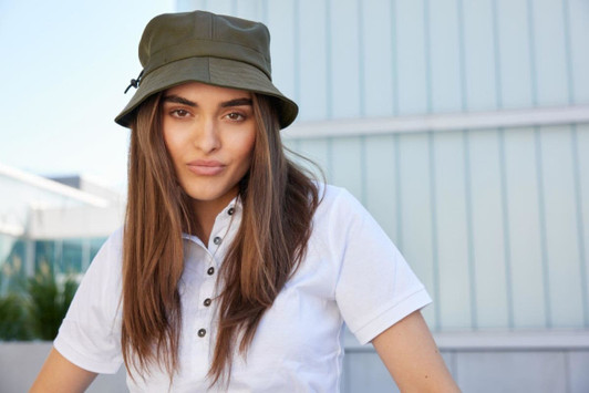 A woman wearing an olive green fisherman hat, with long hair, set against a modern building backdrop.
